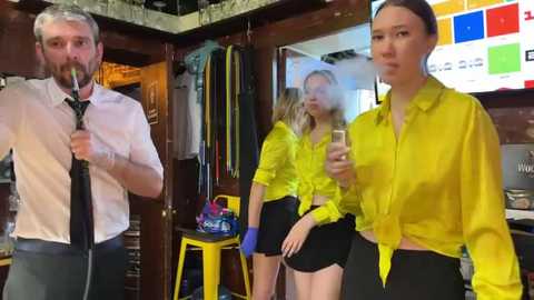 Media: Video of a man and two women in a vibrant yellow room. Man in white shirt, women in yellow shirts and black shorts.