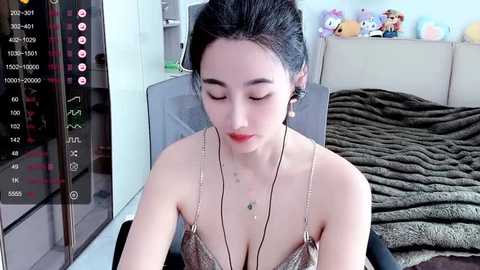 Media: Video of a young Asian woman with dark hair, fair skin, and small breasts, wearing a low-cut metallic top, seated in a modern bedroom with a plush bed, plush toys, and a mirror.