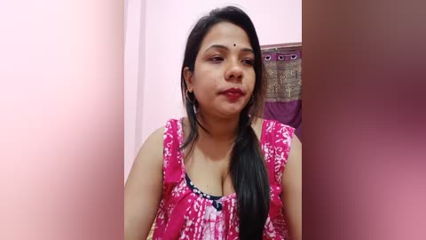 Media: Video of a South Asian woman with medium brown skin, black hair in a side braid, wearing a bright pink sleeveless top with white floral patterns, in a pink-walled room.