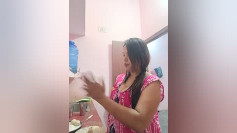 Media: Video of an Asian woman with long black hair in a pink blouse, holding a blue water bottle, standing in a small, brightly lit room with pastel walls.