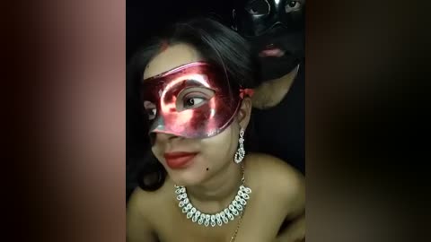 Media: Video of a woman with light brown skin, wearing a red Venetian mask, dangling earrings, and a blingy necklace, with a black background.