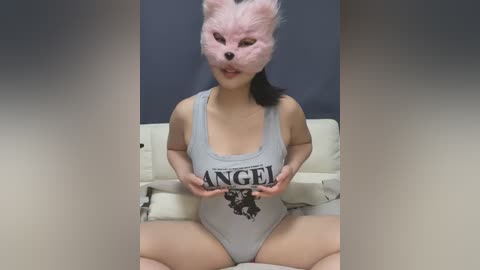 Media: Video of an East Asian woman with fair skin, black hair, wearing a gray Angel Onesie with a wolf mask, seated on a beige couch against a dark background.