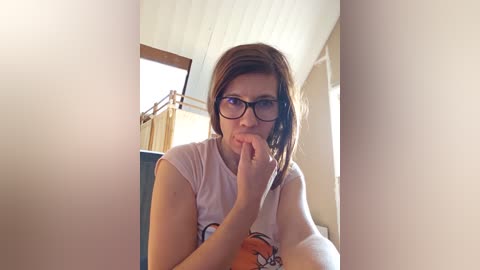 Media: Video of a young Caucasian woman with shoulder-length brown hair, wearing glasses and a white tank top, biting her finger thoughtfully in a dimly lit room with a wooden staircase and cream-colored walls.