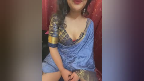 Media: Video of a South Asian woman with medium skin tone, long dark hair, and red lipstick, wearing a blue saree with gold embroidery, sitting on a red draped chair, smiling.