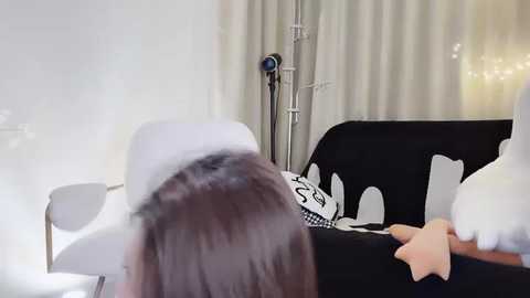 Media: Video of a woman with long, straight hair, reclining on a black sofa, wearing a white headband, in a minimalist room with beige curtains and a medical device in the background.