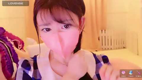 Media: Video of an East Asian woman with fair skin, wearing a blue and white plaid shirt, holding a cloth over her mouth. Background includes a bed, curtains, and a radiator.