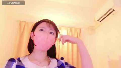 Media: Video of a young Asian woman with straight brown hair, wearing a blue and white striped shirt, blowing into her right hand in a dimly lit room with yellow curtains and an air conditioner.