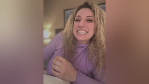 Media: Video of a smiling, light-skinned woman with curly blonde hair, wearing a lavender top, leaning on a bed in a softly lit, beige-walled room.