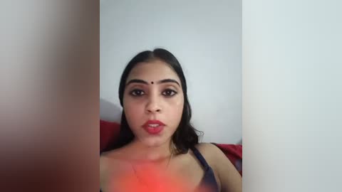 Media: Video of a South Asian woman with fair skin, dark hair, and a bindi, wearing a dark top, sitting against a plain white wall, partially obscured by a red curtain.