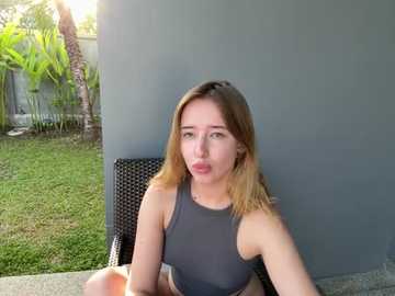 Media: Video of a young Caucasian woman with shoulder-length blonde hair, wearing a grey tank top, sitting outside near a grey wall, making a playful pout.