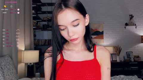 Media: Video of an Asian woman with fair skin, long black hair, wearing a red tank top, standing indoors in a modern room with a white brick wall and wooden furniture.