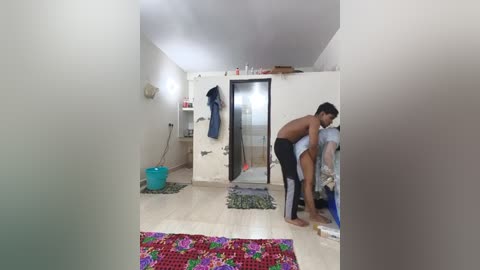 Media: A video of a small, cluttered room with a shirtless man in black shorts bent over a bed, while a woman in a white dress stands next to him. The room has light wood flooring, a red floral rug, and a white wall with a blue bag and clothes hung.