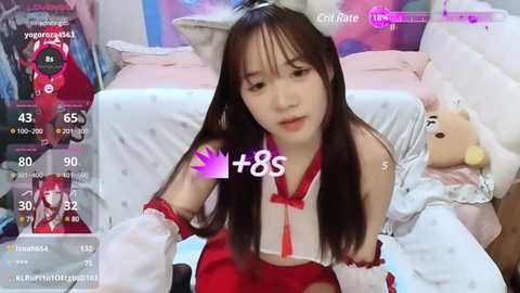 Media: Video of an East Asian woman with long black hair, wearing a red and white maid costume, sitting on a bed with stuffed animals. The background features a soft pink and white room with a TV displaying a virtual character.