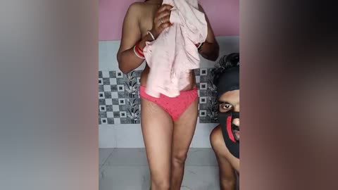 Media: Video of a slender, dark-skinned woman with medium breasts, wearing pink panties and a light pink shirt, holding it up, in a tiled bathroom with checkered wall tiles and a black-haired person in the background.