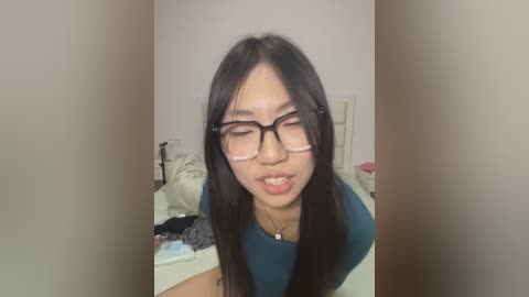 Media: A video of an Asian woman with long black hair and glasses, wearing a teal top, standing in a messy bedroom. She appears to be in her twenties, with a neutral expression.