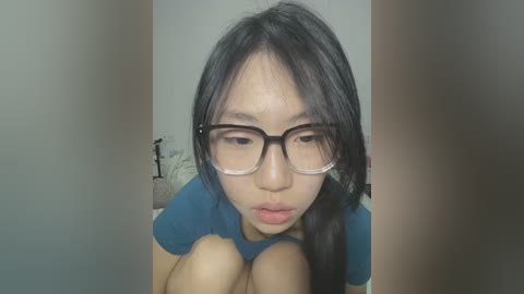 Media: Video of an Asian woman with straight black hair, wearing glasses, a teal shirt, and a neutral expression, sitting on a bed with white sheets, blurred background.