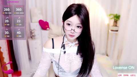 Media: Video of an East Asian woman with long black hair, wearing glasses, a white lace top, and headphones, in a softly lit room with a pink pillow and potted plant.