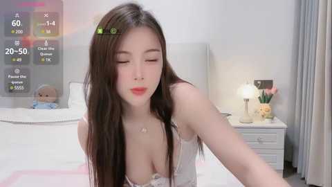 Media: A young Asian woman with long brown hair, wearing a white lace bra, leans forward in a modern bedroom with a white bed, nightstand, and a digital clock.