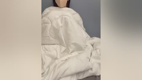 Media: A video of a person with light skin and dark hair wrapped in white bedding, partially obscured by a grey wall. The bedding appears soft and slightly wrinkled.