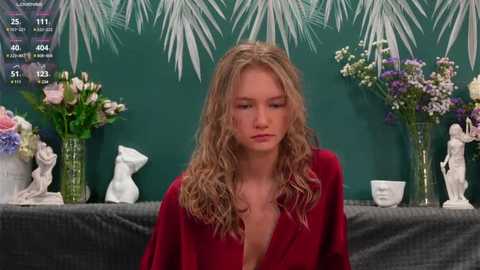 Media: Video of a blonde woman with curly hair, wearing a red blouse, sitting in front of a green wall adorned with floral decorations and statuettes.