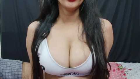 Media: Video of a woman with long black hair, wearing a white sports bra that emphasizes her large, natural breasts. She has fair skin, and the background features a dark curtain and a pink floral pillow.