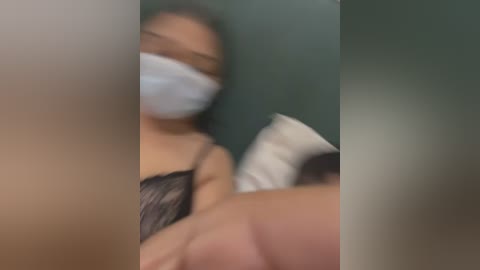 Media: A blurry video shows a person with light brown skin wearing a black lace bra and a white face mask, lying on a bed with white pillows and a green wall in the background.