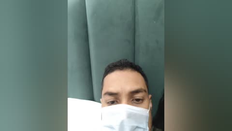 Media: A video of a man wearing a surgical mask, lying on a green cushioned surface, possibly in a medical setting, with a blurred background.