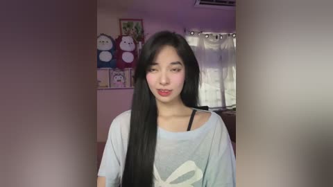 Media: Video of an Asian woman with long black hair, wearing a loose gray t-shirt with a white graphic, in a dimly lit room adorned with stuffed animals and a window.