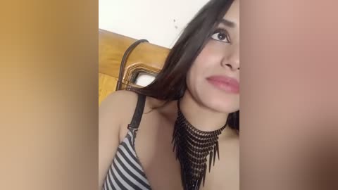 Media: Video of a young woman with medium skin tone and long dark hair, wearing a black and white striped bra and a layered black beaded necklace. She has a subtle smile, and the background features a wooden chair and white wall.