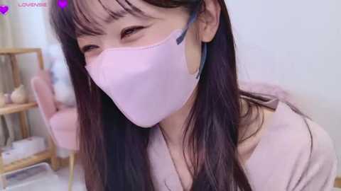 Media: A video of an Asian woman with long black hair, wearing a pink face mask, winking, and smiling, indoors with a wooden chair and a shelf in the background.