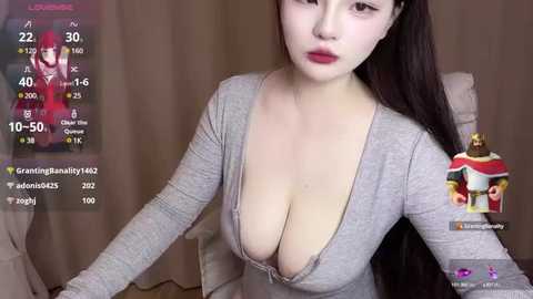 Media: Video of an East Asian woman with long black hair, wearing a low-cut gray top revealing ample cleavage, sitting in a beige chair. Background shows a live streaming platform with user stats and a profile picture.