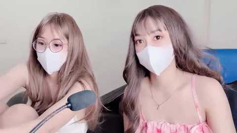 Media: Video of two young Asian women with long brown hair, wearing white masks and pink tank tops, seated closely, one using a microphone, indoors.