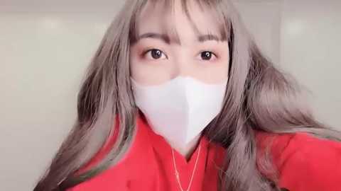 Media: A close-up video of a young Asian woman with long, wavy gray hair and a white surgical mask covering her nose and mouth. She wears a bright red jacket and has fair skin. The background is a plain, light-colored wall.