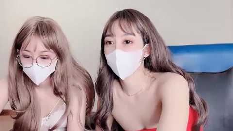 Media: Video of two East Asian women with long, straight brown hair, wearing white face masks and red strapless tops, seated closely together, indoors, with a blue chair and beige wall background.