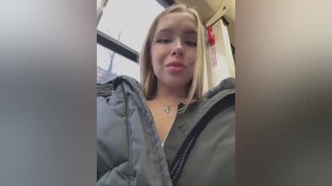 Media: A video of a young Caucasian woman with light skin, straight blonde hair, and wearing a gray jacket, taken from a low angle inside a bus.