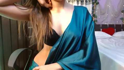 Media: Video of a young woman with long, straight brown hair, wearing a black bra and teal satin robe, standing in a dimly lit bedroom with a white bed and draped curtains.