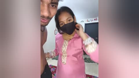 Media: Video of a young woman in a pink kurta with floral embroidery, wearing a black mask, standing next to a man in a shirt, in a brightly lit room with a TV in the background.