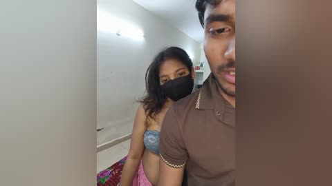 Media: A video of an Asian woman in a blue bra, mask, and colorful skirt, standing next to a man in a brown shirt, both indoors.