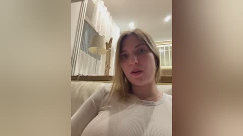 Media: Video of a Caucasian woman with long blonde hair, wearing a white ribbed sweater, sitting indoors. Background features beige walls, white curtains, and a window.