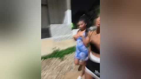 Media: Video of a young girl, wearing a blue swimsuit, running on a path with green grass, next to a woman. Background shows a building and concrete path.