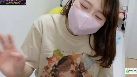 Media: A video of an East Asian woman with straight brown hair, wearing a pink face mask, beige T-shirt with a cartoon character, and glasses, waving in a brightly lit room with stuffed animals in the background.