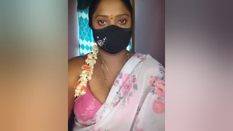 Media: Video of a South Asian woman with medium brown skin wearing a pink floral saree, black mask, and flower garland, against a blue and white diamond-patterned backdrop.