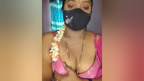 Media: Video of a South Asian woman with medium brown skin, wearing a black face mask, pink lace bra, gold necklace, and a floral garland.