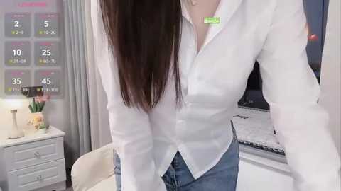 Media: Video of a woman with long dark hair in a white blouse and blue jeans, standing in a modern office with a calendar, desk, and plants.