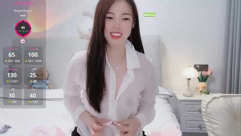 Media: A video of an East Asian woman with long black hair, wearing a white blouse, sitting on a bed with white linens, smiling. The room features a white headboard, a nightstand with a lamp, and a window with light curtains.