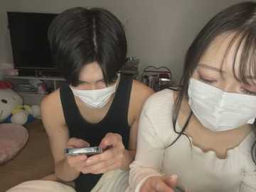 Media: Video of two East Asian women in masks, one with short black hair and a black tank top, the other with long black hair and a white shirt, playing with a phone, in a cluttered living room.