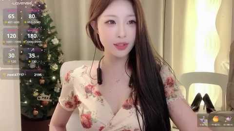 Media: Video of an East Asian woman with long, straight brown hair, wearing a floral blouse, sitting in a cozy living room with a Christmas tree in the background.