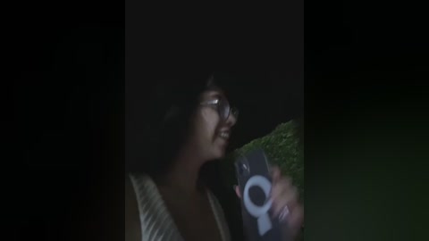 Media: Video of a smiling woman with glasses, wearing a light-colored top, taking a selfie against a dark background with a green bush.