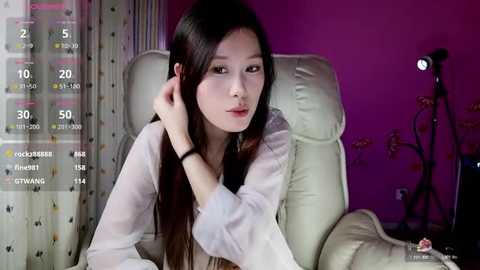 Media: Video of an East Asian woman with long black hair, wearing a white blouse, sitting on a white chair in a dimly lit room with purple walls.