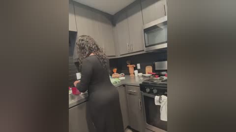 Media: A video of a woman with curly hair, wearing a black dress, cooking in a modern kitchen with light-colored cabinets, black countertops, and appliances.